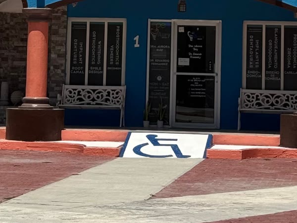 La entrada de la clínica en la planta baja de Plaza Rincones de México: fachada azul con el número 1, una puerta de vidrio entre dos ventanas rotuladas con nombres de tratamientos y, delante, dos escalones y una rampa con el símbolo de silla de ruedas pintado.